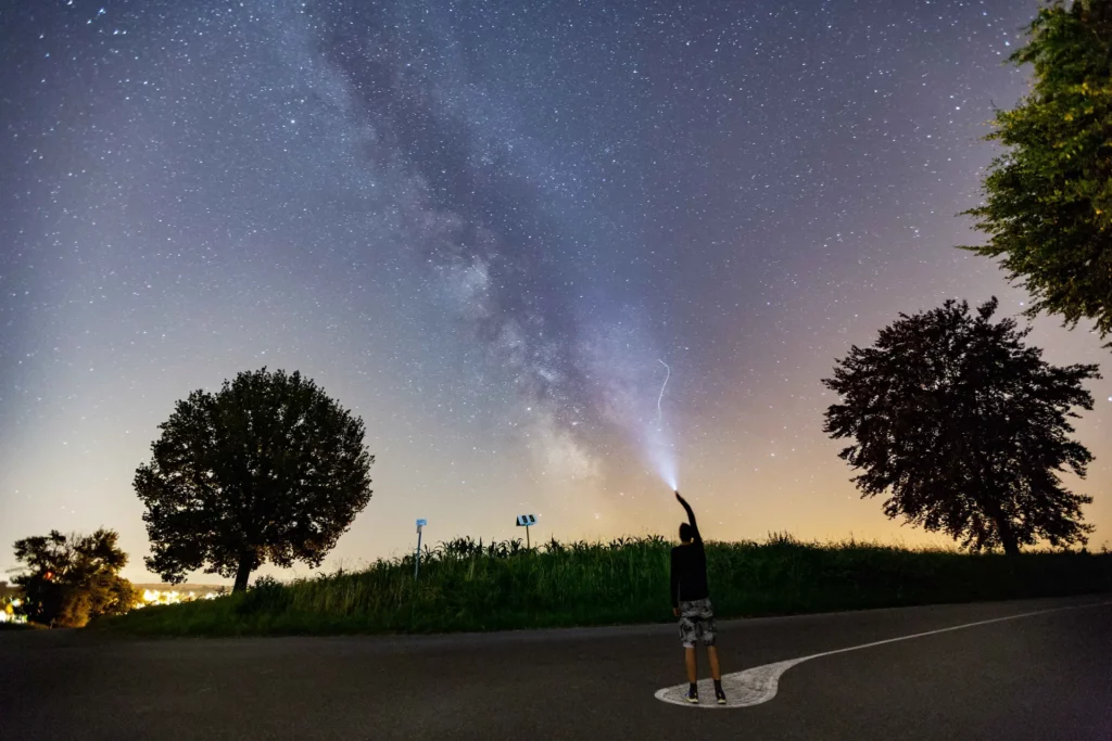 Voie lactée en Alsace – Nuit étoilée capturée par Tijoe, photographe à Mulhouse Un homme éclaire la Voie lactée avec une lampe torche sous un ciel étoilé, photo nocturne en Alsace