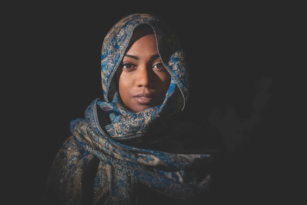 Portrait clair-obscur – Femme en voile oriental sous lumière dramatique Portrait artistique d’une femme en voile bleu doré, éclairée par un faisceau de lumière sur fond noir