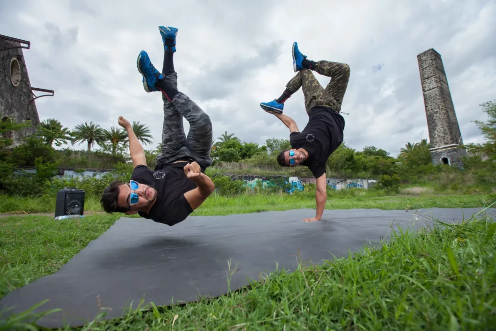 Deux danseurs de breakdance exécutant des figures acrobatiques en extérieur sur un tapis noir, devant une ancienne cheminée industrielle à La Réunion