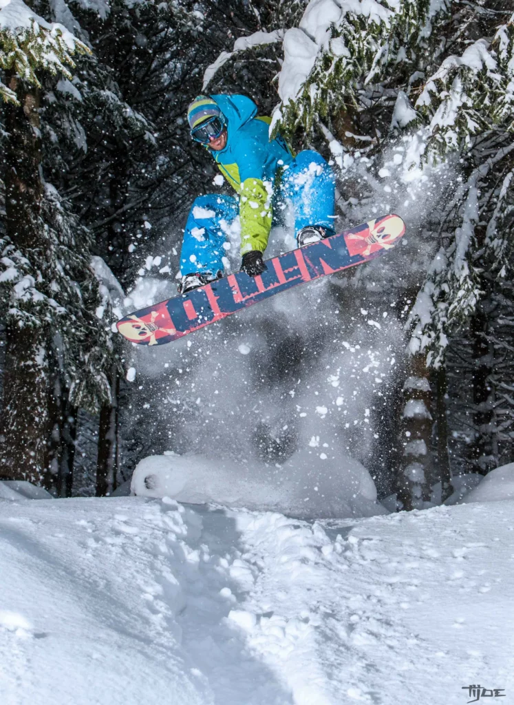Snowboarder effectuant un saut dans une forêt enneigée, éclaboussant la poudreuse en plein vol