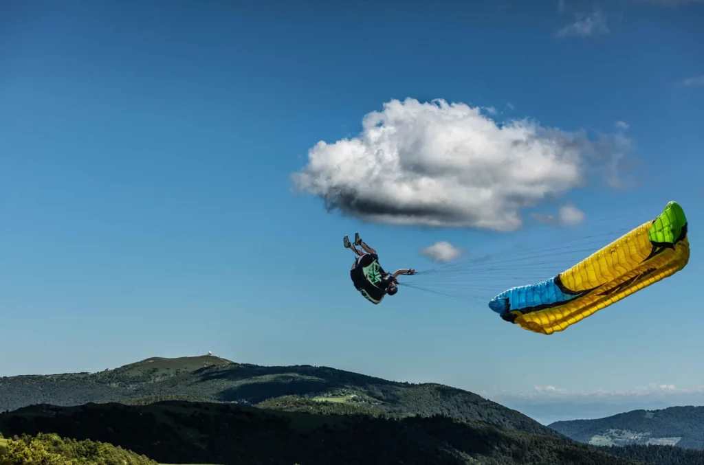 Parapentiste réalisant une figure acrobatique en vol devant des nuages au-dessus d’un paysage de collines verdoyantes